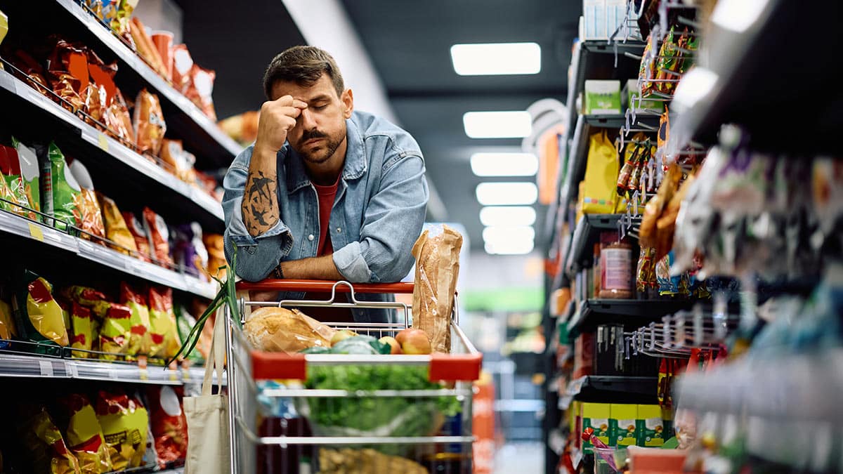 Frustrierter Mann im Geschäft mit Lebensmitteln im Einkaufswagen