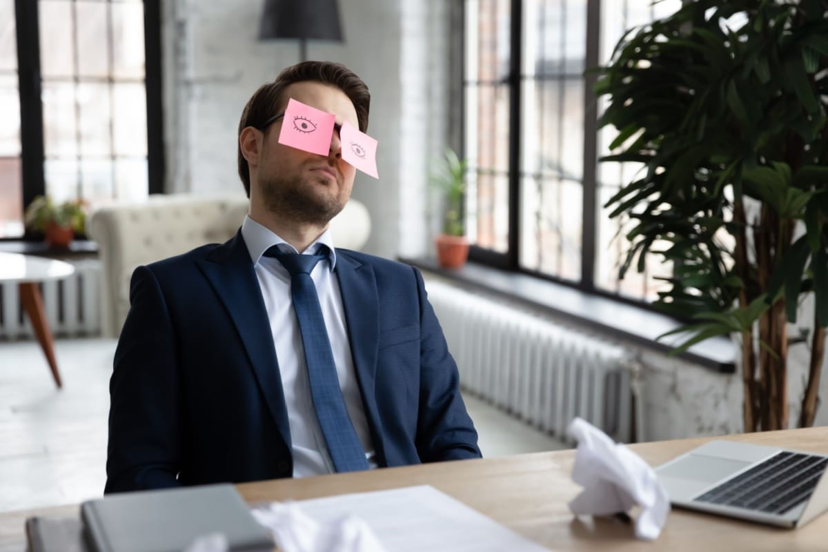 Gelangweilter Arbeitnehmer im Anzug sitzt mit Post-its auf den Augen am Schreibtisch – Symbol für Unzufriedenheit im Job