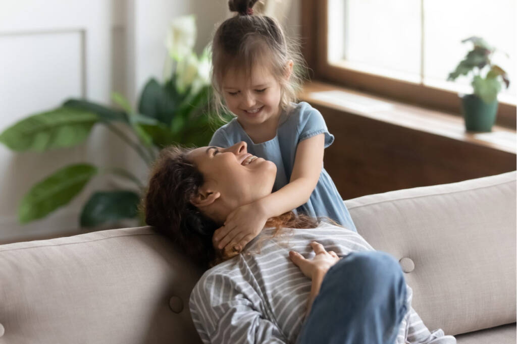 Alleinerziehende Mutter spielt lachend mit Tochter auf dem Sofa