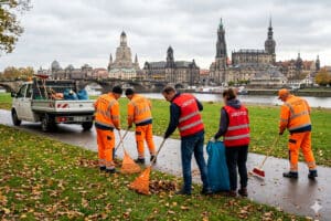 Drei städtische Mitarbeiter in orangefarbener Warnkleidung und zwei Personen in roten Westen mit der Aufschrift „JOBCENTER“ kehren gemeinsam Laub am Elbufer vor der Silhouette der Dresdner Altstadt.