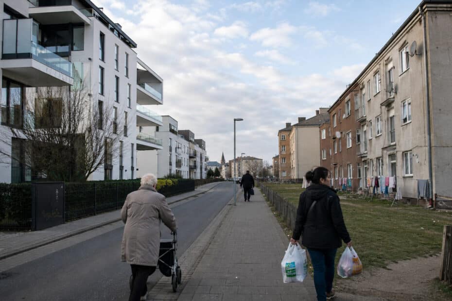 Ältere Frau mit Rollator und Passantin mit Einkaufstaschen in einer Straße zwischen modernen und einfachen Wohnhäusern als Symbol für soziale Ungleichheit und Armutsgefährdung in Deutschland