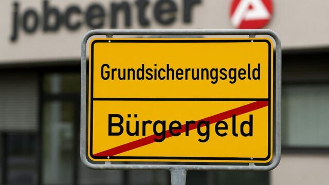 Schild mit Grundsicherungsgeld statt Bürgergeld vor unscharfem Jobcenter-Gebäude