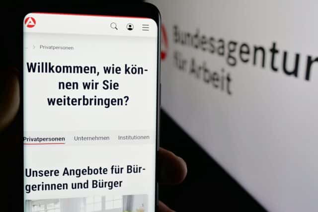 Mobiltelefon ruft Bundesagentur für Arbeit Seite auf vor Monitor mit BA-Seite