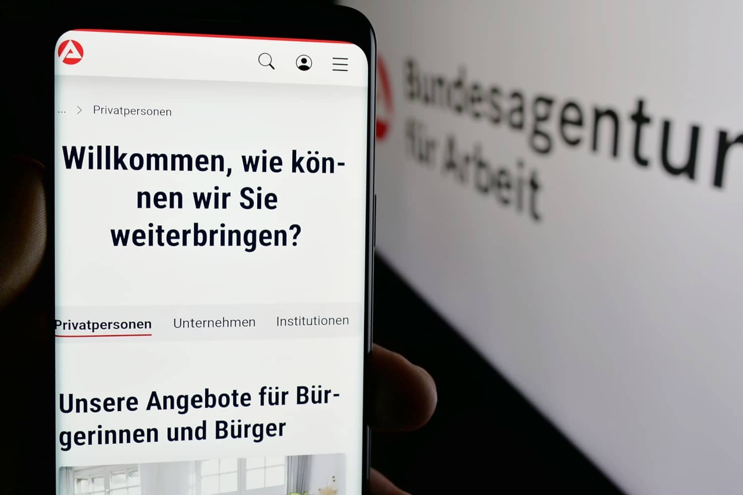 Mobiltelefon ruft Bundesagentur für Arbeit Seite auf vor Monitor mit BA-Seite