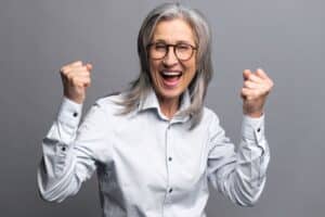 Frau mit Brille jubelt mit erhobenen Fäusten und breitem Lächeln, Symbolbild für Beamten-Vollkasko und volle Bezüge trotz Krankheit.