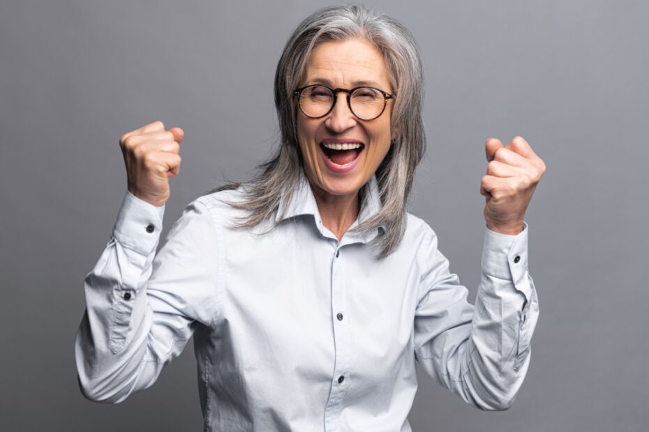 Frau mit Brille jubelt mit erhobenen Fäusten und breitem Lächeln, Symbolbild für Beamten-Vollkasko und volle Bezüge trotz Krankheit.