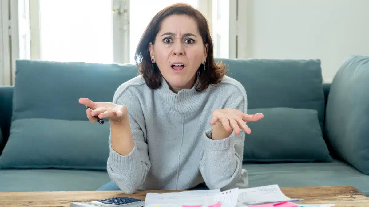 Frustrierte Frau mit Taschenrechner und Papieren am Tisch. Sie drückt Verwirrung oder Ärger über finanzielle Probleme und Bürokratie (Jobcenter/Bürgergeld-Bescheide) aus.