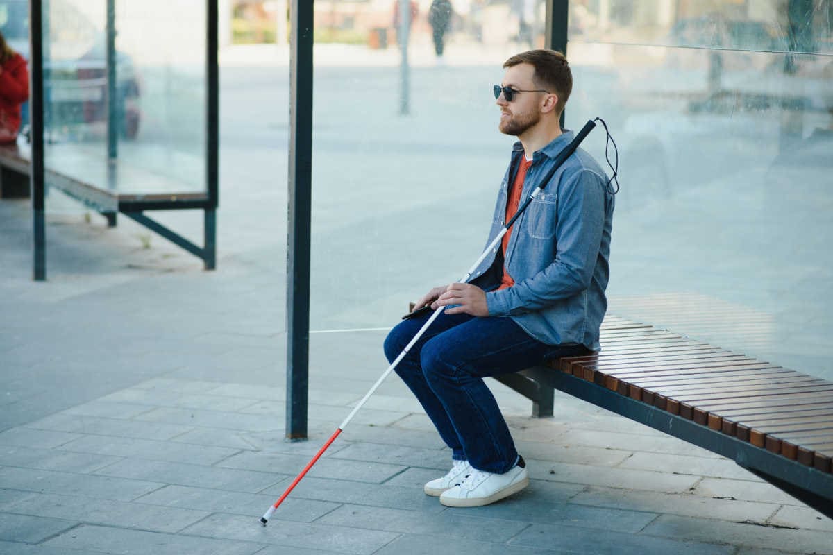 blinder Mann sitzt mit Stock an Bushaltestelle