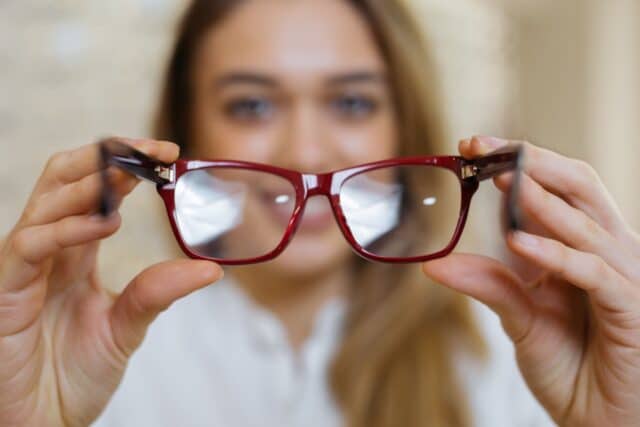 Nahaufnahme einer Brille mit rotem Gestell, die von einer leicht unscharfen Frau im Hintergrund präsentiert wird; symbolisiert Sehhilfe und Reparaturkosten.