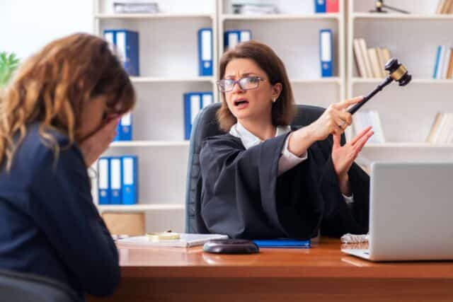 Richterin hält Hammer und weist mit abweisender Geste in Richtung Tür, signalisisert Ablehnung. Vor ihr sitzt eine Person mit gesenktem Kopf und Händen im Gesicht.
