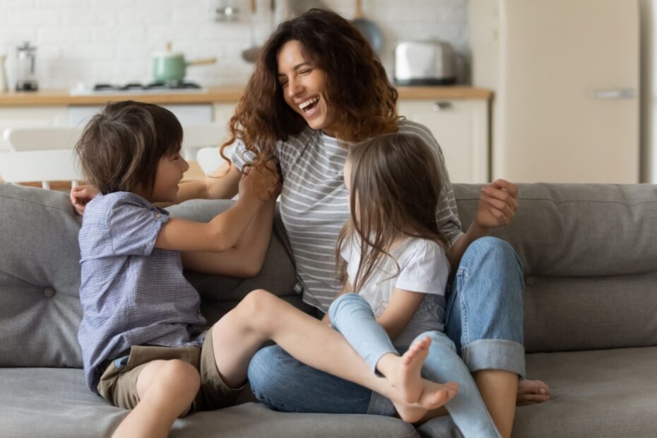 Lachende alleinerziehende Mutter spielt mit ihren zwei Kindern im Wohnzimmer.