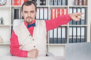 Strenger Jobcenter-Mitarbeiter oder Sachbearbeiter mit Fliege sitzt am Schreibtisch und weist mit dem Finger auf die Tür.