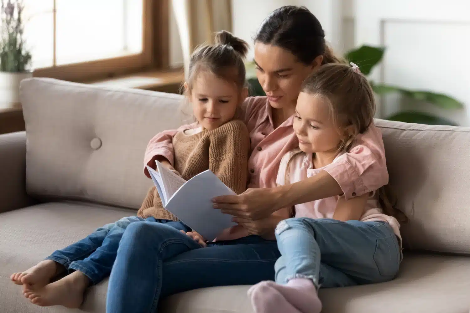 Alleinerziehde Mutter sitzt mit ihren Kindern auf dem Sofa und liest ein Buch.