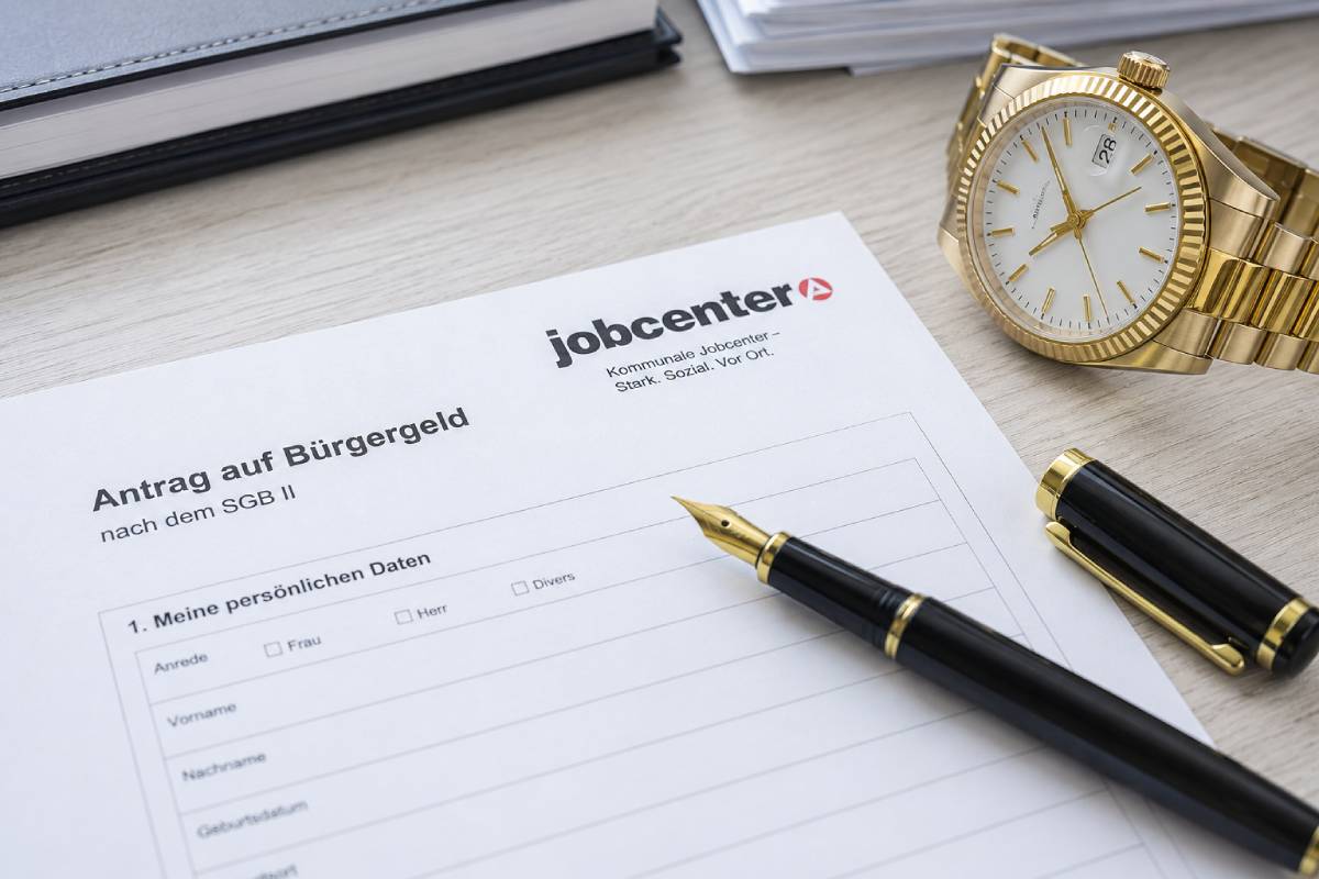 Ein Bürgergeld-Antrag des Jobcenters auf einem Schreibtisch, daneben liegen ein edler schwarzer Füllfederhalter und eine hochwertige goldene Armbanduhr.