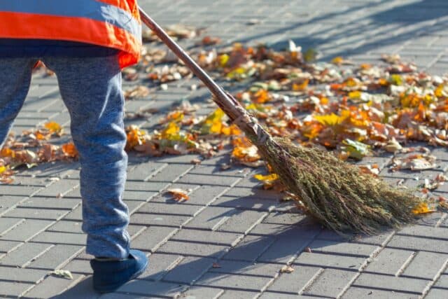 Eine Person in orangefarbener Warnweste kehrt mit einem Besen Herbstlaub auf Gehweg. Tätigkeit symbolisiert gemeinnützige Arbeiten im Rahmen der Bürgergeld Arbeitspflicht