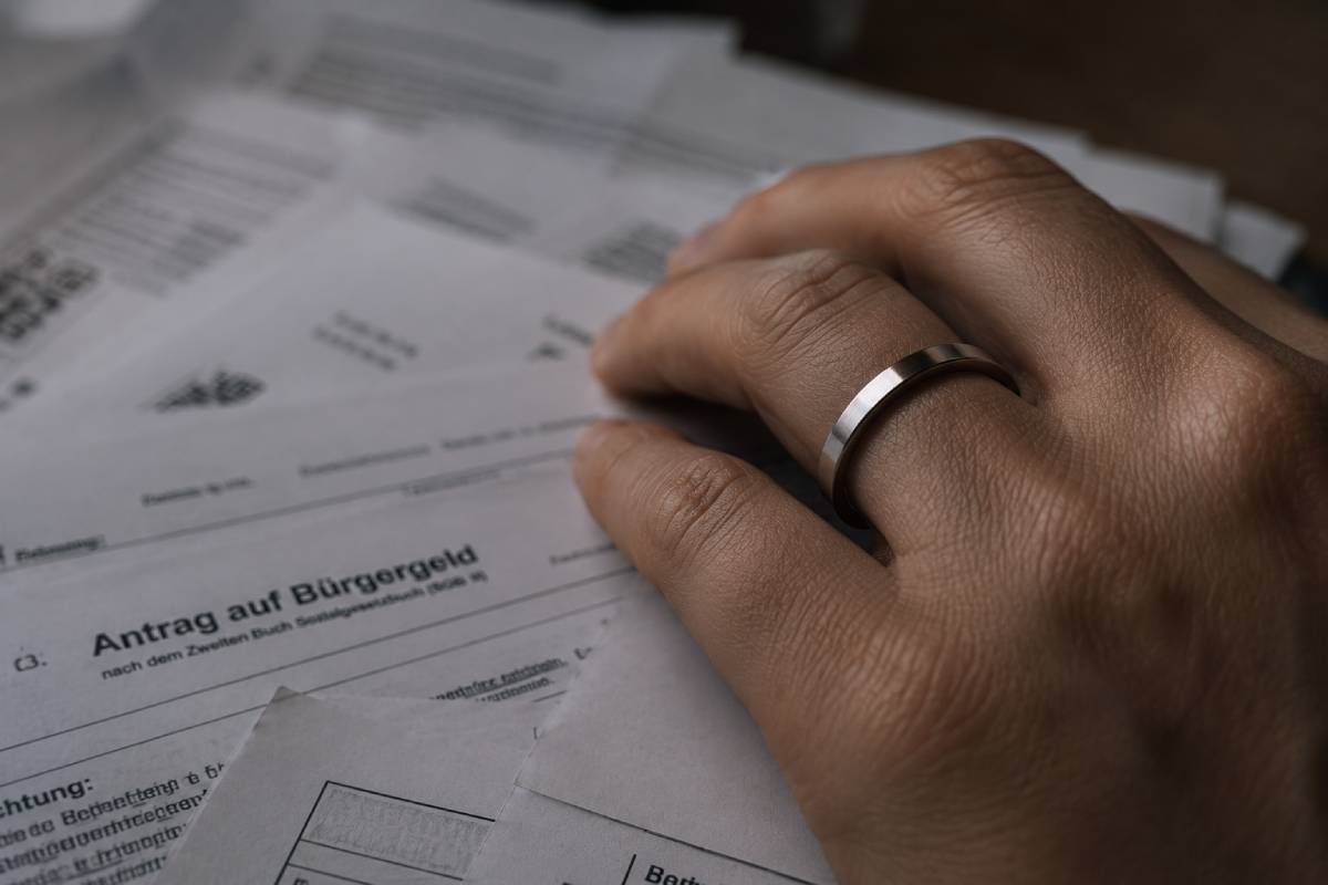 Nahaufnahme einer Hand mit Ehering, die auf verschiedenen amtlichen Bürgergeld-Formularen liegt.