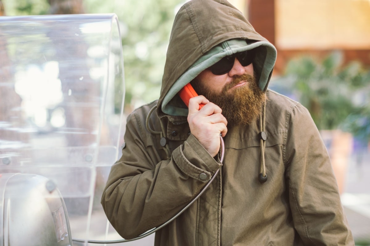 Mann mit Kapuze und Sonnenbrille telefoniert an öffentlicher Telefonzelle – Symbolbild für anonyme Hinweise oder Denunziation im Zusammenhang mit Bürgergeld und Jobcenter.