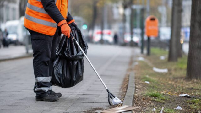Eine Person in Warnweste sammelt mit einem Greifwerkzeug Müll auf einem Gehweg.