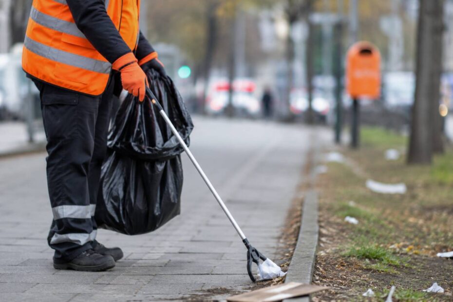 Eine Person in Warnweste sammelt mit einem Greifwerkzeug Müll auf einem Gehweg.