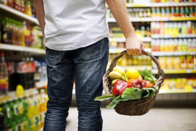 Mann steht im Supermarkt, in der Hand ein Einkaufskorb voll Obst und Gemüse. Symbolisch für Mehrbedarf bei chronischen Krankheiten