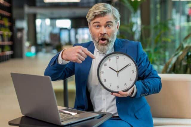 Mann zeigt auf Uhr zur Verdeutlichung einer ablaufenden Bürgergeld-Frist am Laptop.