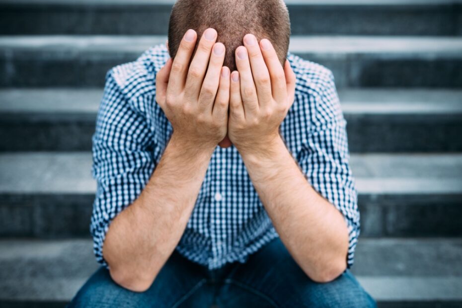 Junger Mann mit kariertem Hemd sitzt auf einer Treppe, das Gesicht in den Händen vergraben. Symbolisiert Frustration und Überforderung nach Fehlern im Bürgergeld-Verfahren.