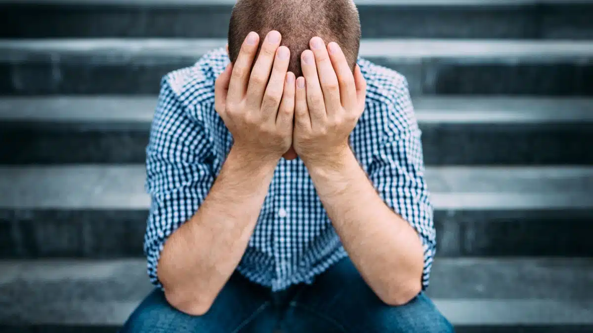 Junger Mann mit kariertem Hemd sitzt auf einer Treppe, das Gesicht in den Händen vergraben. Symbolisiert Frustration und Überforderung nach Fehlern im Bürgergeld-Verfahren.