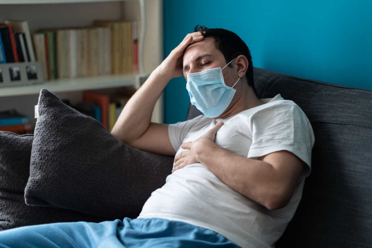 Mann krank mit Maske auf Sofa sitzend