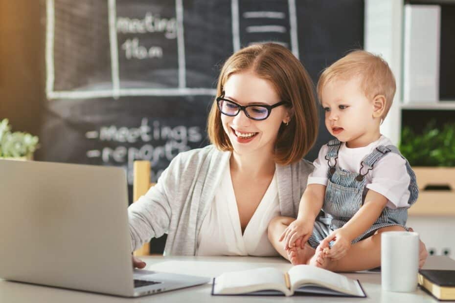 Junge Mutter arbeitet am Laptop, ihr Baby sitzt daneben auf dem Schreibtisch