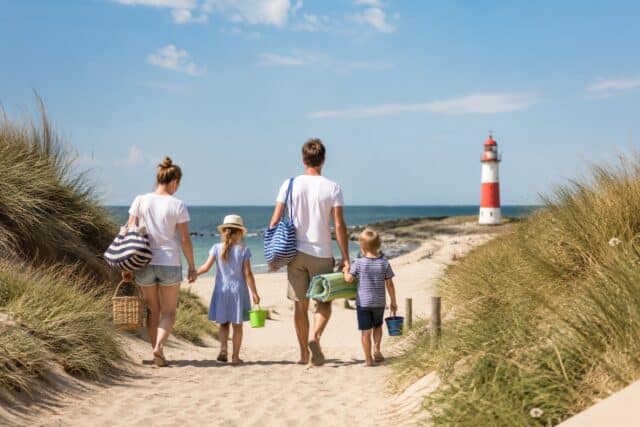 Familenurlaub. Eltern gehen mit zwei Kindern zum Strand, auf dem ein rot-weißer Leuchtturm steht