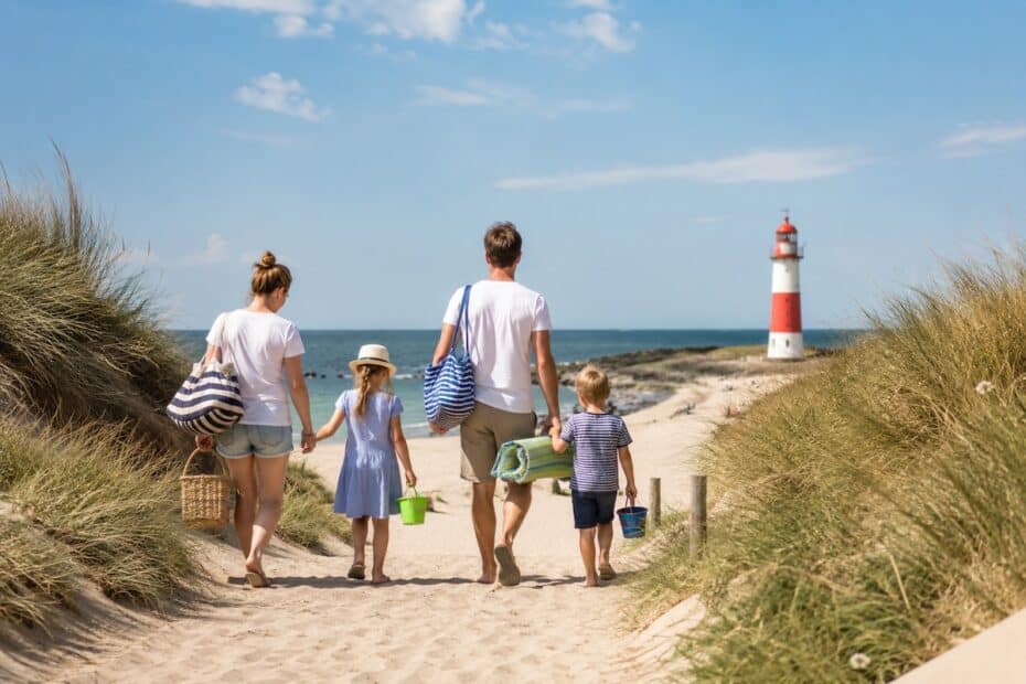 Familenurlaub. Eltern gehen mit zwei Kindern zum Strand, auf dem ein rot-weißer Leuchtturm steht