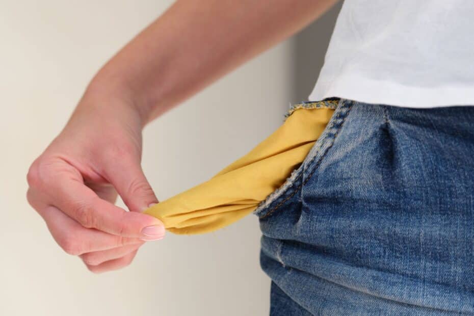 Text: Nahaufnahme einer Hand, die eine leere, nach außen gezogene gelbe Hosentasche zeigt, um Mittellosigkeit oder Geldmangel zu symbolisieren.