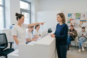 Frau steht mir ihrer Gesundheitskarte in der Hand am Tresen beim Arzt und wird von der MFA abgewiesen.