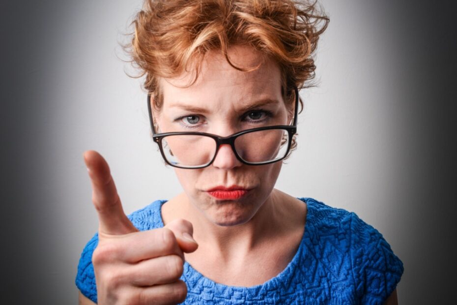 Eine streng blickende Frau mit Brille zeigt mahnend mit dem Zeigefinger auf den Betrachter, symbolisiert Kontrolle und Strenge.