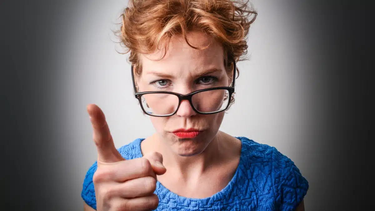 Eine streng blickende Frau mit Brille zeigt mahnend mit dem Zeigefinger auf den Betrachter, symbolisiert Kontrolle und Strenge.