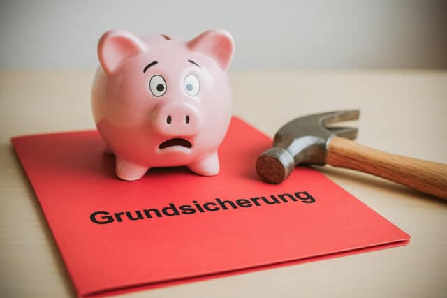 Erschrockenes Sparschwein neben Hammer auf roter Mappe mit Aufschrift "Grundsicherung"