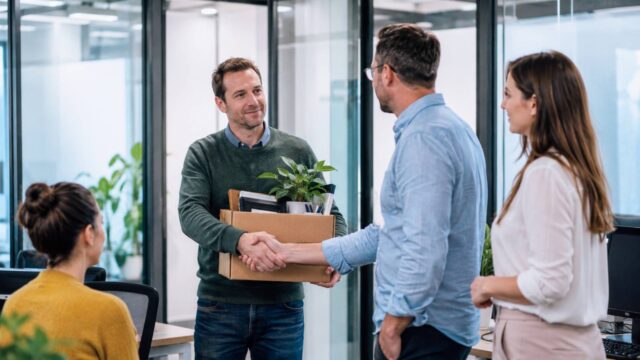Mann mit Karton voller persönlicher Gegenstände verabschiedet sich von seinen Kollegen im Büro.