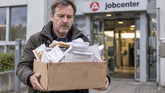 Mann trägt einen überfüllten Karton mit ungeordneten Belegen vor ein Jobcenter.