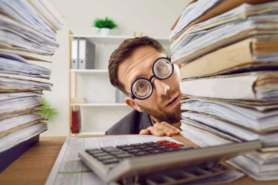 Mann mit runder Brille blickt erschöpft zwischen zwei hohen Aktenstapeln auf einen Schreibtisch mit Taschenrechner – Sinnbild für Bürokratie und stockende Integration im Bürgergeld‑System