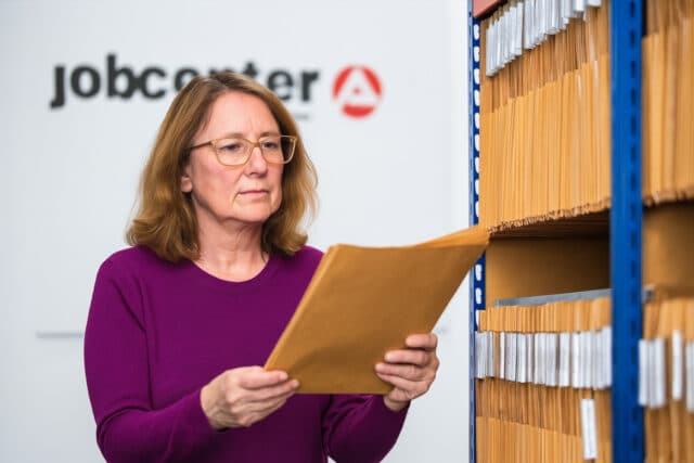Frau mittleren Alters steht im Archiv des Jobcenters vor Regalen mit zahlreichen Akten. Im Hintergrund ist das unscharfe Jobcenter-Logo an der Wand zu erkennen.