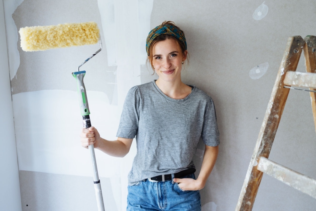 Lachende Frau mit Stirnband und Malerrolle vor Leiter und weißer, gespachtelter Wand (Symbol für Renovierung in Eigenleistung)