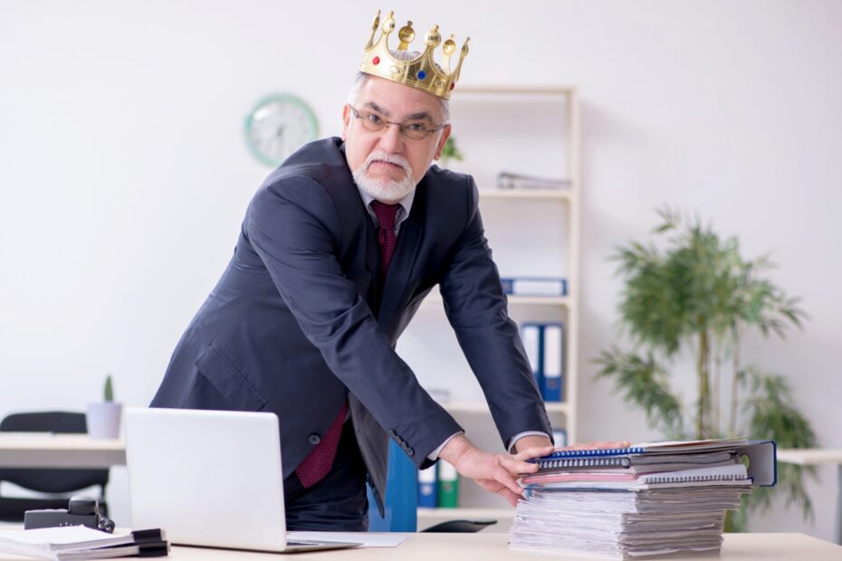 Büroangestellter steht mit aufgesetzter Krone hinter dem Schreibtisch