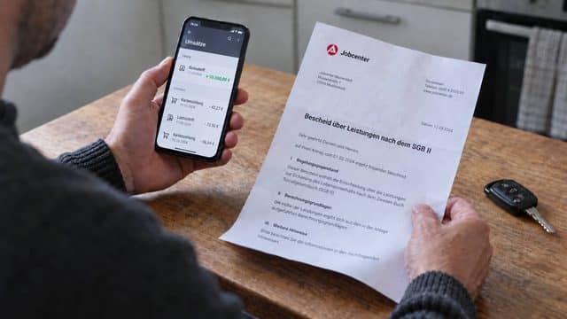 Mann prüft eine Überweisung über 10.000 Euro und einen Jobcenter-Bescheid am Küchentisch