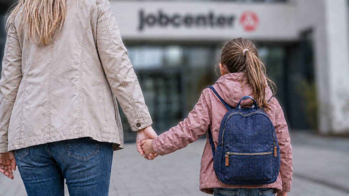 Mutter geht mit ihrer Tochter an der Hand auf den Eingang eines Jobcenters zu.