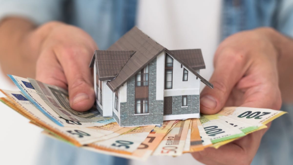 Hände halten ein Muster-Eigenheim auf Euro-Banknoten als Baufinanzierung