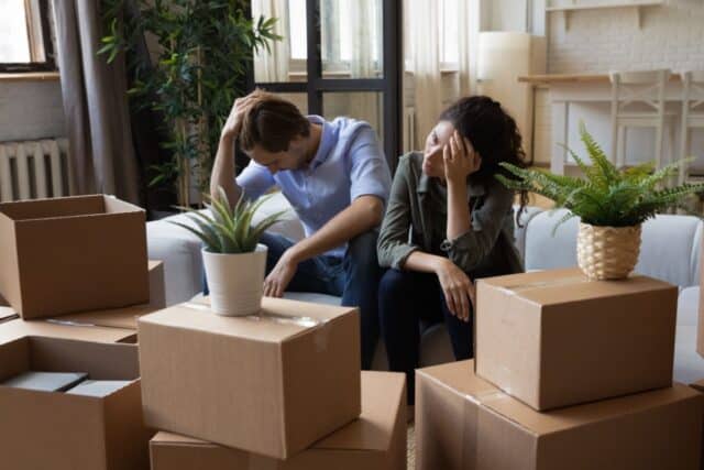 Trauriges oder verzweifeltes junges Paar sitzt umgeben von Umzugskartons auf dem Sofa. Symbolbild für Kündigung, Wohnungsverlust und Mietschulden.