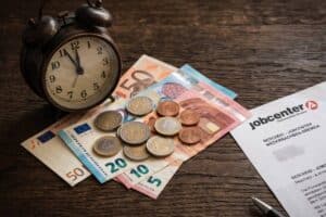 Stapel Euro-Scheine und Münzen neben einer tickenden Uhr und einem Jobcenter-Schreiben zur Aufrechnung eines Darlehens vom Bürgergeld.