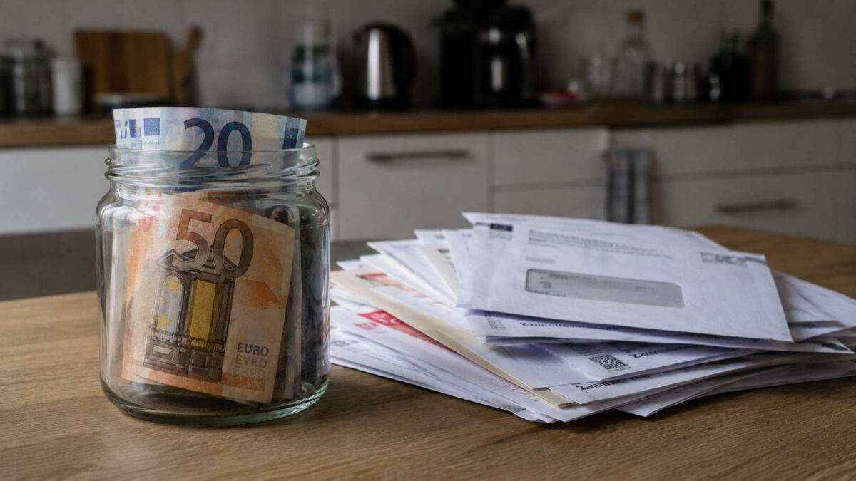 Glas mit Euro-Banknoten neben einem Stapel unbezahlter Rechnungen und Mahnungen auf einem Küchentisch