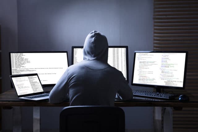 Hacker in Kapuze sitzt vor mehreren Monitoren