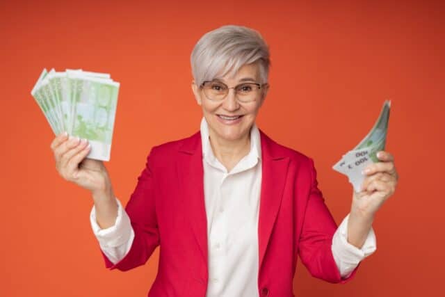 Lachende ältere Frau mit kurzem, hellen Haar und Brille in einem knallroten Blazer, die in jeder Hand Bündel von 200-Euro-Scheinen hält, vor einem orangen Hintergrund. Die Szene illustriert eine "üppige Rente" oder hohe Geldgewinne.